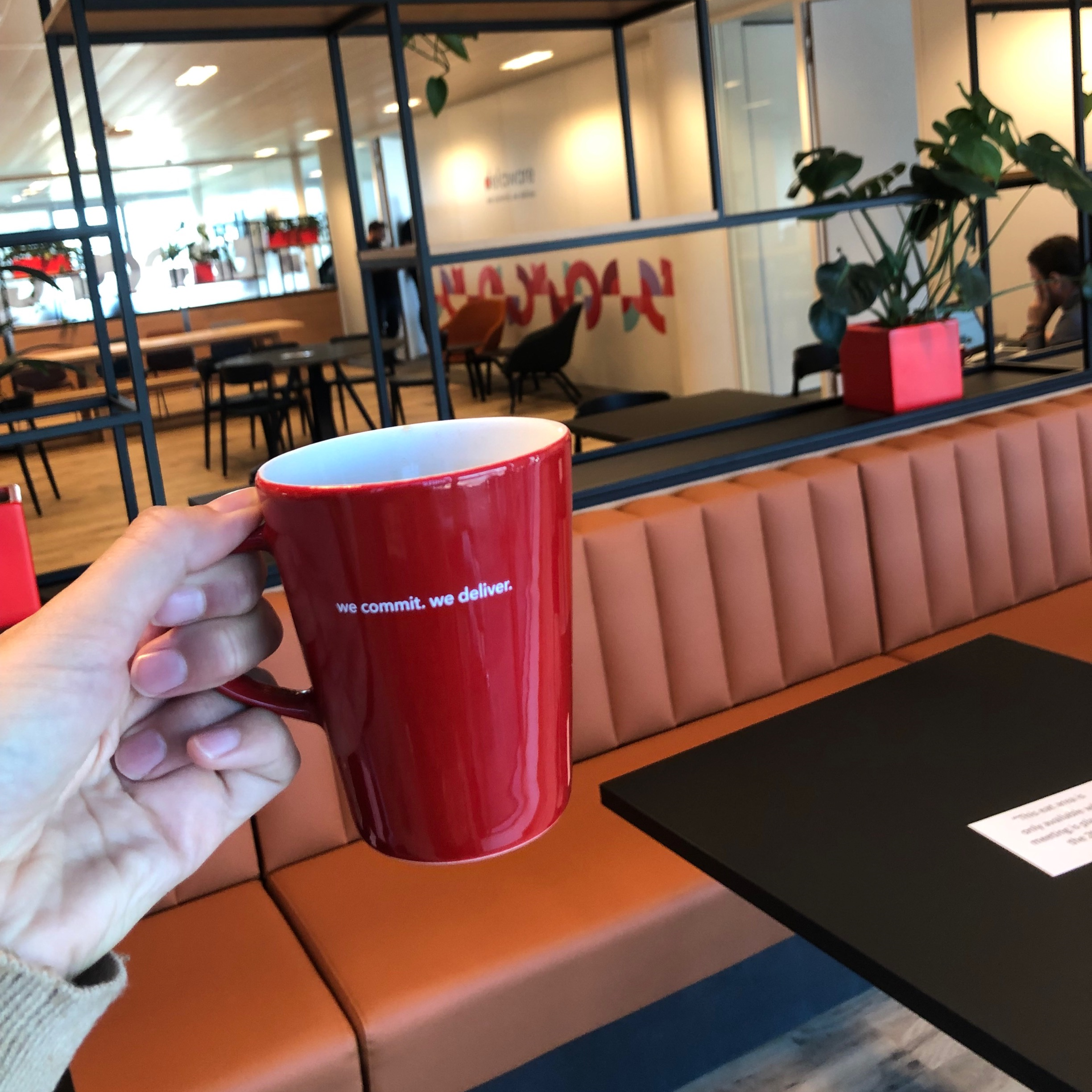A hand is holding a cup with the we commit. we deliver. tagline in one of the Belgian offices of delaware