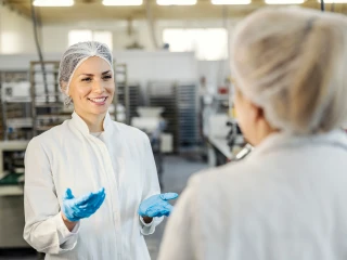 picture of 2 women wearing protective white coats and hair nets chatting in a food production facility